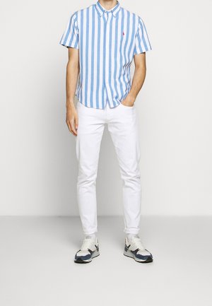Camisa de manga corta con botones, de rayas verticales azules y blancas, con un logo rojo en el pecho, combinada con pantalones blancos y zapatillas multicolores.