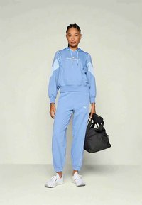 Kvinne iført lyseblå hettegenser og joggebukser, hvite joggesko, holder en svart treningsveske, står mot en ensfarget lys bakgrunn.