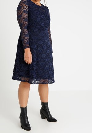 Femme portant une robe en dentelle bleu marine au genou avec des manches longues et des bottines noires à talons, debout sur un fond blanc.