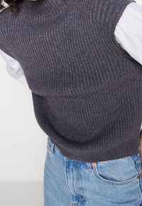 Grauer, gerippter ärmelloser Pullover aus weichem Material mit hohem Kragen. Kombiniert mit hellblauen, hoch taillierten Jeans.