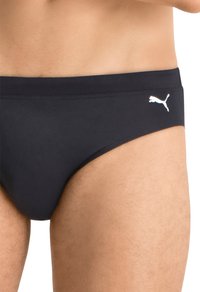 Svarte svømmebukser med en jevn overflate, med en hvit Puma-logo på siden og en lav midje for minimal dekning.