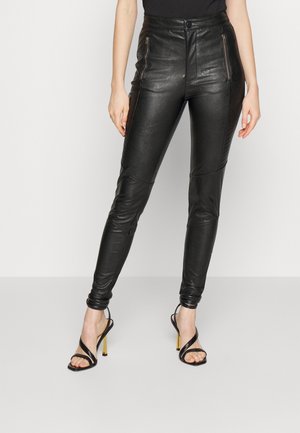 Kobieta ubrana w czarne, skórzane spodnie z wysokim stanem, z przodu zamki błyskawiczne przy kieszeniach, oraz czarne, na wysokim obcasie, sandały na paski, stojąca na tle jednolitego tła.