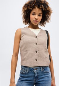 Νεαρή γυναίκα με σγουρά μαλλιά, φοράει μπεζ γιλέκο με κουμπιά πάνω από λευκό τοπ και μπλε τζιν, κρατώντας μια μαύρη τσάντα ώμου.