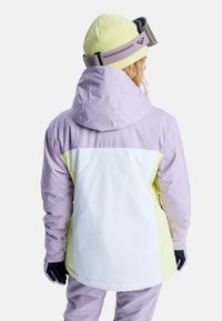Fioletowa i żółta kurtka narciarska z białym panelem z tyłu, z kapturem i czarnymi rękawicami, noszona z jasną żółtą czapką i goglami.