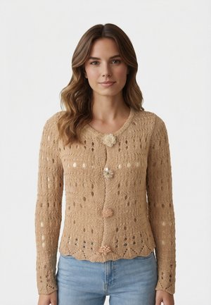 Donna con capelli castano ondulati, indossa un cardigan beige lavorato a maglia con bottoni decorati a fiore e jeans azzurri, in piedi su uno sfondo bianco.