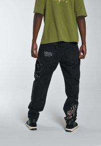 Jeans de mezclilla negra con diseños bordados de tigre y pantera en la parte trasera, con un corte relajado y cierres de botón en los laterales. Llevados con una camisa de color verde oliva.