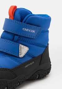 Botte imperméable bleue avec deux bandes velcro, accent argenté et semelle texturée noire. Comprend un talon renforcé et une languette orange.