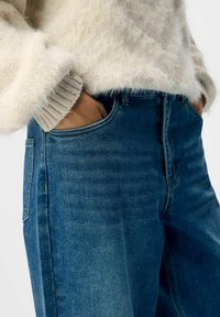 Person som bär blå denimjeans och en fluffig benvit tröja med händerna i framfickorna.