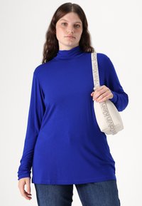 Long-sleeve turtleneck shirt in solid royal blue fabric, paired with a cream-colored handbag featuring textured accents and a patterned strap.