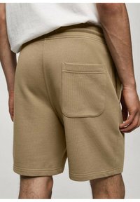 Homme portant un short beige avec une poche arrière, se tenant devant un fond uni, visible de la taille au mi-thigh.