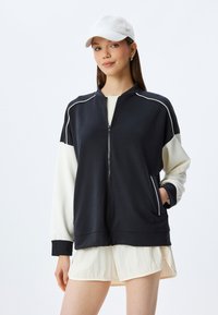 Svart och gräddfärgad zip-up jacka med hög krage, kontrasterande ärmar, sidofickor och vita piping-detaljer. Mjuk tygkänsla.