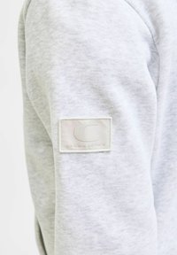 Sudadera gris claro con una tela texturizada. Presenta un parche rectangular crema en la manga, grabado con "C" y "JACK & JONES."
