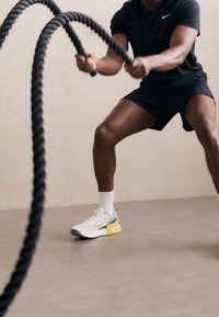 Camisa corta negra de manga corta y pantalones cortos, zapatos deportivos grises y amarillos, agarrando gruesas cuerdas de batalla negras, de pie sobre un suelo liso.
