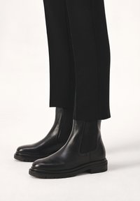 Black leather ankle boots with elastic side panels, round toe, and flat rubber sole, worn with black tailored pants. Smooth texture and sleek design.