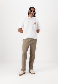 White short-sleeve t-shirt with red graphic, beige textured pants, and beige shoes with dark accents. Black round sunglasses.