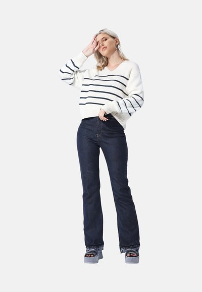 Maglione a righe bianche e navy con scollo a V, vestibilità ampia; jeans a zampa d'elefante di colore blu scuro; sandali con plateau e suola testurizzata, con le dita dei piedi scoperte.