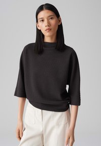 Haut en tricot texturé noir avec un col montant et des manches trois-quart, associé à un pantalon de couleur claire. Coupe lisse et structurée.