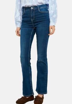 Kobieta ubrana w ciemnoniebieskie dżinsy o prostych nogawkach i wysokim stanie, jasnoniebieską bluzkę z długim rękawem w kwiatowy wzór oraz brązowe mokasyny.