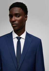 Completo blu navy in tessuto texturizzato, design slim-fit, rever a punta e una tasca sul petto. Abbinato a una camicia bianca e una cravatta scura.