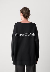 Črna oversize sv sweatshirt iz mehke tkanine, s širokim ovratnikom in izstopajočim belim logotipom "Marc O'Polo" na hrbtu.