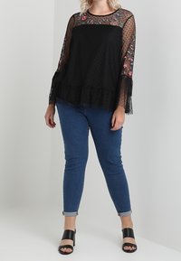 Haut transparent à pois noirs avec broderie florale, ourlet à volants superposés ; associé à un jean skinny bleu et des sandales à talons hauts noires.
