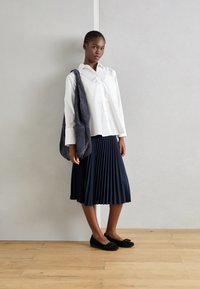 Camicia bianca con bottoni, gonna plissettata blu navy, borsa in suede grigia, ballerine nere con dettaglio a fiocco. Texture morbide e design classico.
