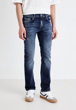 Osoba ubrana w dopasowane niebieskie dżinsy z delikatnymi przetarciami oraz białe trampki z czarnymi paskami, stojąca na jednolitym tle.