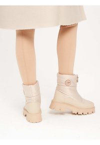 Botas de tobillo beige con parte superior acolchada, punta redondeada y suela gruesa. Presenta un cierre lateral y un acento de logo circular en el eje.