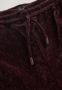 Burgunderfarbene Samtjogginghosen mit einem elastischen Bund, verstellbarem Kordelzug und strukturiertem Stoff für ein angenehmes Tragegefühl. Metallenden an der Kordel.