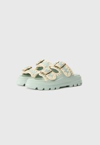 Green sandal with textured straps, featuring cream-colored braided accents and metal buckles. Chunky sole with a tread pattern.