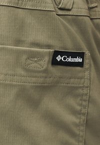 Ετικέτα της μάρκας Columbia στην τσέπη από ελιά πράσινο υφασμα με υφή, που δείχνει λεπτομερείς ραφές σε εξωτερικό ένδυμα.