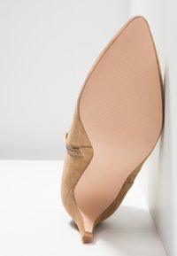 Chaussure beige en daim à talon haut et bout pointu avec fermeture éclair latérale, montrée de la semelle et du talon sur fond blanc.