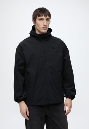 Jeune homme aux cheveux bruns et raides porte une veste noire à capuche zippée et un pantalon sombre, debout devant un fond uni et clair.