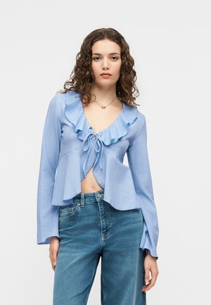 Mladá žena s kučeravými vlasmi, oblečená v svetlomodrom blúzkovom topánke s volánikmi na dlhých rukávoch, previazanom vpredu, a v modrých rifle s vysokým pásom, stojaca na bielom pozadí.