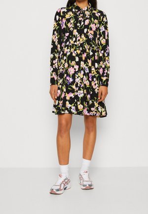 Robe noire à manches longues, imprimé floral en rose et violet, jupe plissée, portée avec des chaussures de sport grises et roses et des chaussettes blanches.