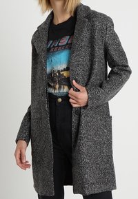 Grijze lange jas van een wollen mix met een reverkraag, sluiting met één knoop en zijzakken, gedragen over een grafisch T-shirt en een broek met hoge taille.