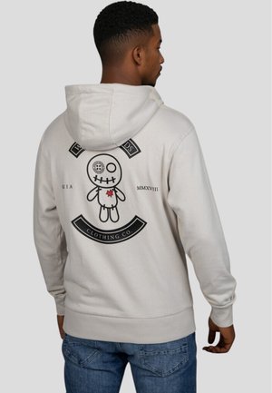 Sudadera gris claro con un gráfico negro de una muñeca cosida en la parte trasera, que cuenta con un detalle en forma de corazón y el texto "CLTHING CO." debajo.