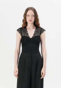 Black bodysuit with lace cap sleeves, deep V-neckline, textured lace bodice, and solid fabric waist. Paired with high-waisted black trousers.