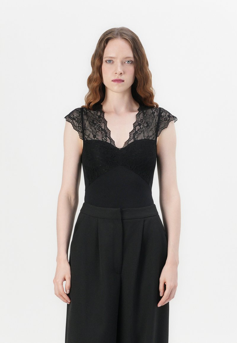 Black bodysuit with lace cap sleeves, deep V-neckline, textured lace bodice, and solid fabric waist. Paired with high-waisted black trousers.