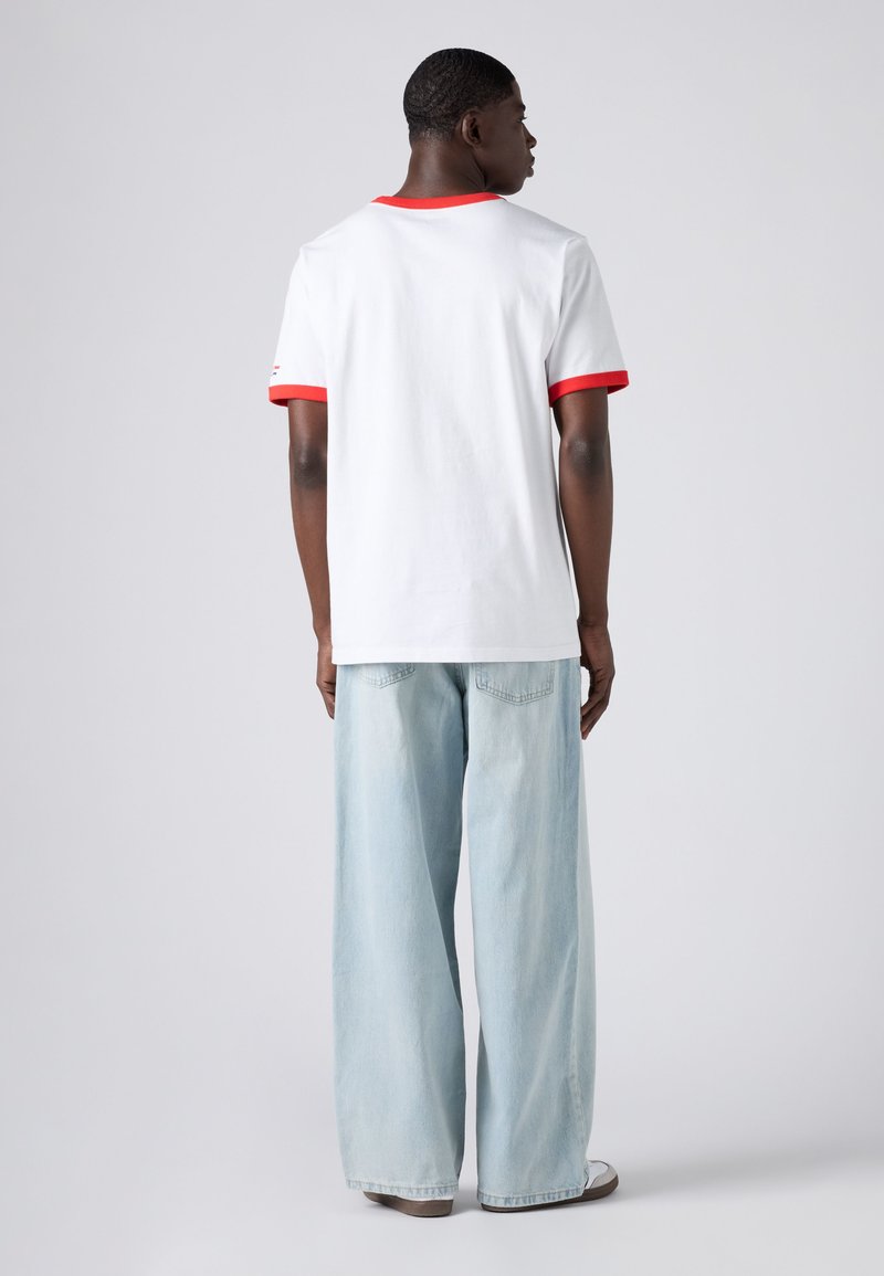 Man facing away wearing white T-shirt with red trim and light blue wide-leg jeans on plain light background.