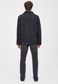 Veste en denim noir avec un col à revers, tissu texturé, boutons en accent, et détail en forme de yoke à l'arrière, associée à un jean foncé et des baskets blanches.