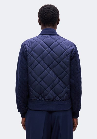 Personne vue de dos portant une veste matelassée bleu marine avec des manches à motif torsadé tricoté et un col et des poignets côtelés.