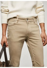 Jeans de algodón beige con un corte ajustado, costuras visibles, bolsillos delanteros y un botón de metal en la cintura. Complementados con un suéter de color claro.