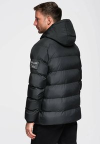 Schwarze Steppjacke mit Kapuze, mit gesteppter Gestaltung, elastischen Bündchen und einem Aufnäher am linken Ärmel. Glattes, glänzendes Material.