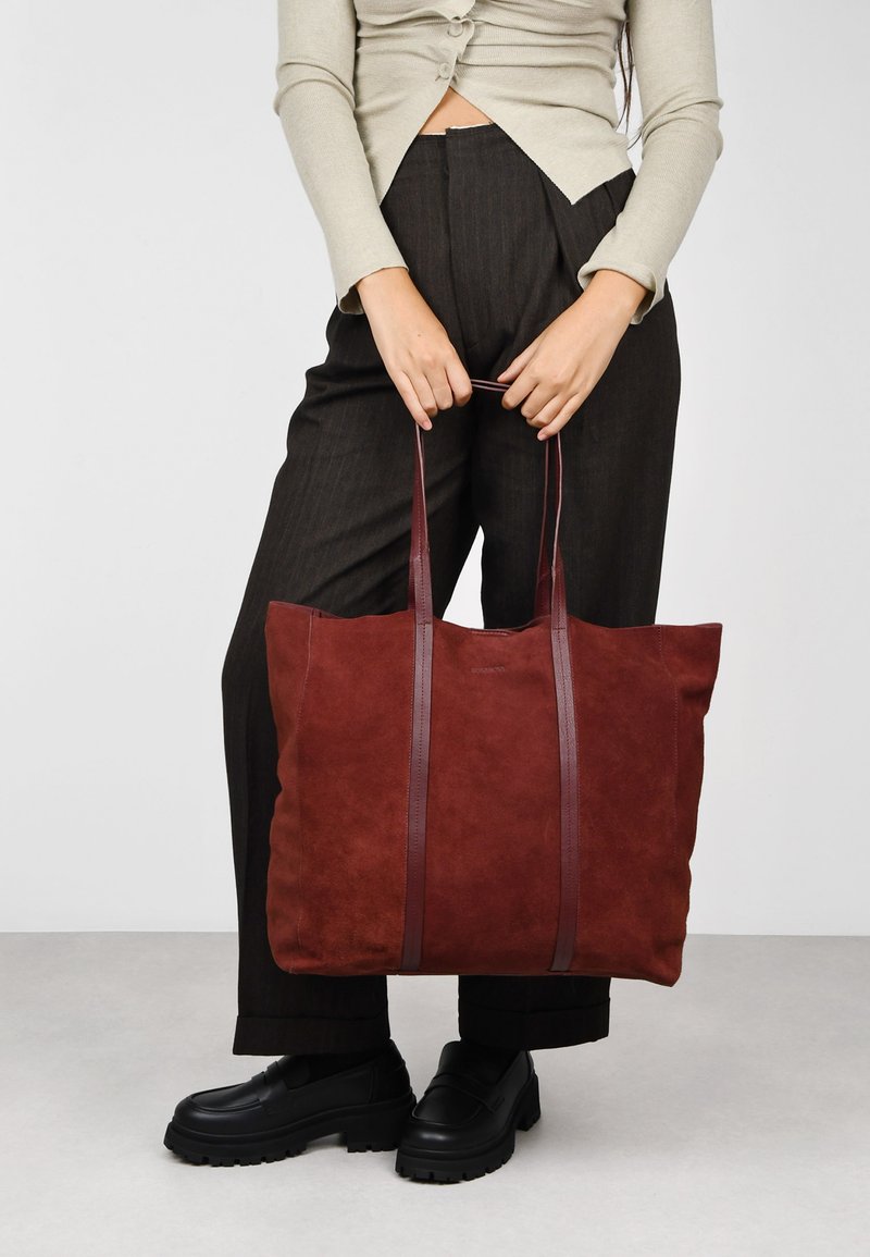 Bolso tote de ante marrón con dos asas de cuero oscuro; forma cuadrada con una textura suave; presenta detalles de costura a lo largo de los laterales.