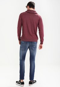 Mörkröd huvtröja i mjukt material med ribbad nederkant, kombinerad med blå denimjeans och svarta sneakers, sedd bakifrån.