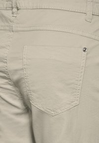 Beige bomullbyxor med en fuskficka på baksidan prydd med en metallknapp, stickade sömmar och en slät yta.