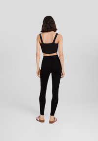 Mustat ribattut crop-topit leveillä olkaimilla yhdistettynä vyötärön ylle ulottuviin mustiin leggings-housuihin. Mallilla on jalassaan liukuhihna-sandaalit, joissa on vaaleanpunaiset hihnat ja ruskea pohja.