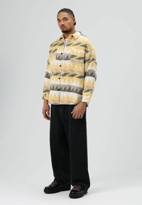 Overhemd met knoopsluiting, bedrukt met abstracte geometrische vormen in geel, grijs en groen. Zwarte jeans met wijde pijpen en zwarte schoenen met roze veters.