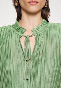 Grüne plissierte Bluse mit rüschigem Kragen, Schlüssellochöffnung mit Bindebändern und metallischen Akzenten für einen verstellbaren Verschluss. Weicher, strukturierter Stoff.